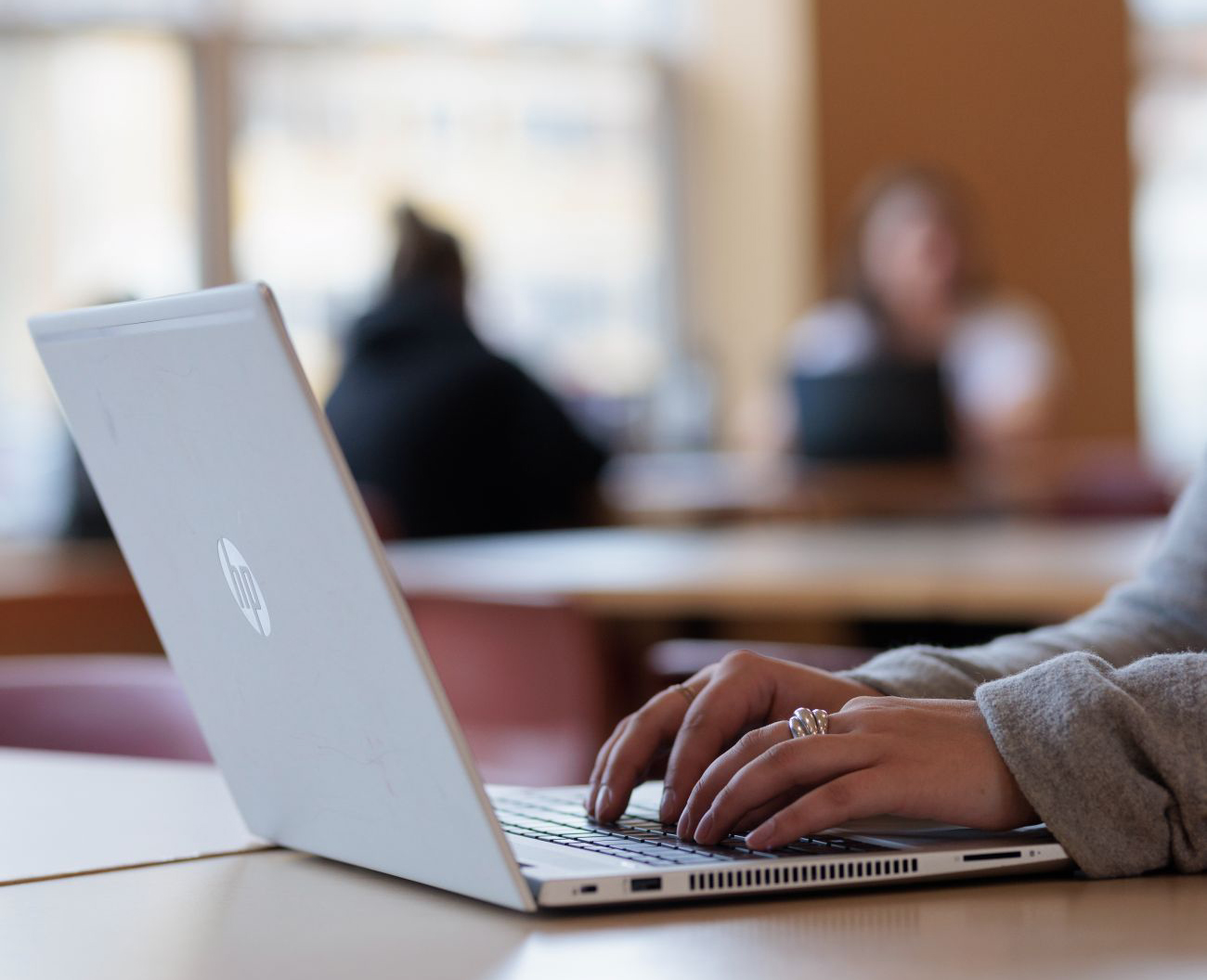 A close-up, shallow-focus shot of a person’s hands typing on a silver HP laptop in a bright, indoor public space.