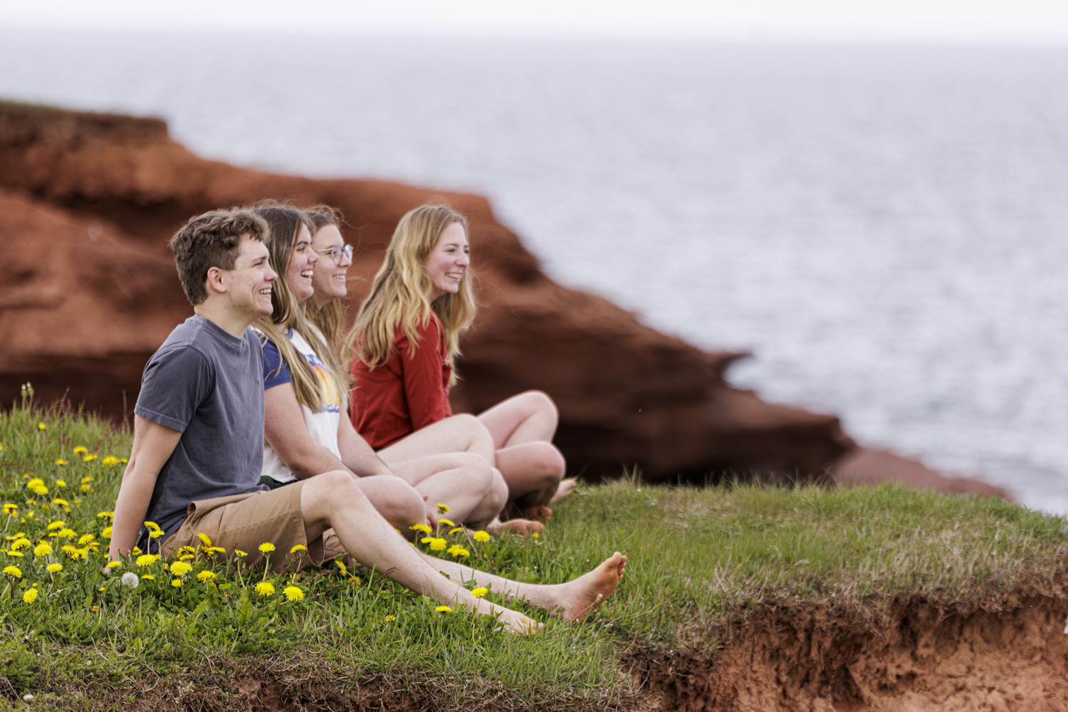 Four young adults sit on a grassy, red-earth cliffside, smiling and looking out over the ocean.