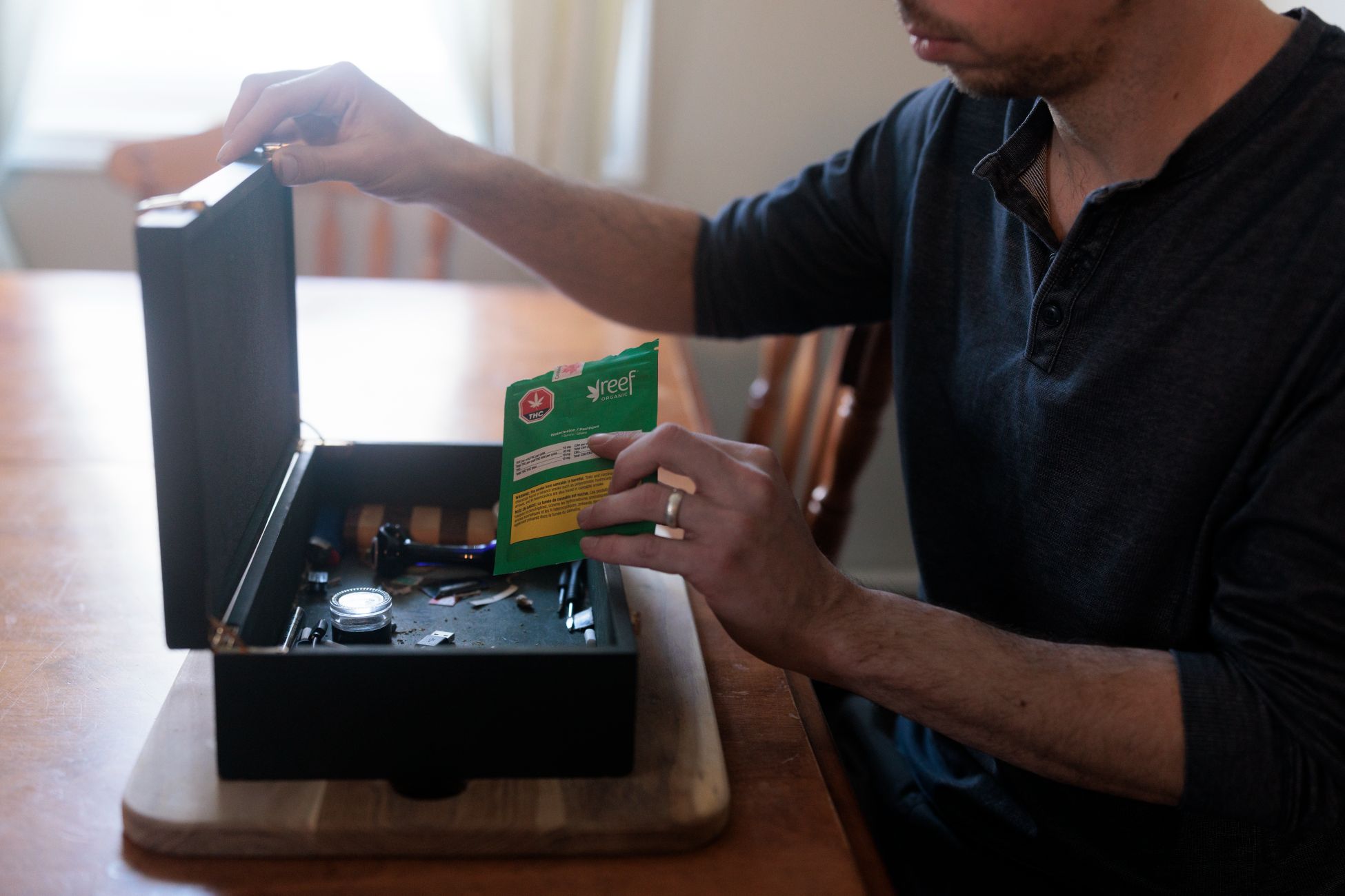 A person sitting at a wooden table, opening a small black storage box and holding a green package of Reef brand cannabis.